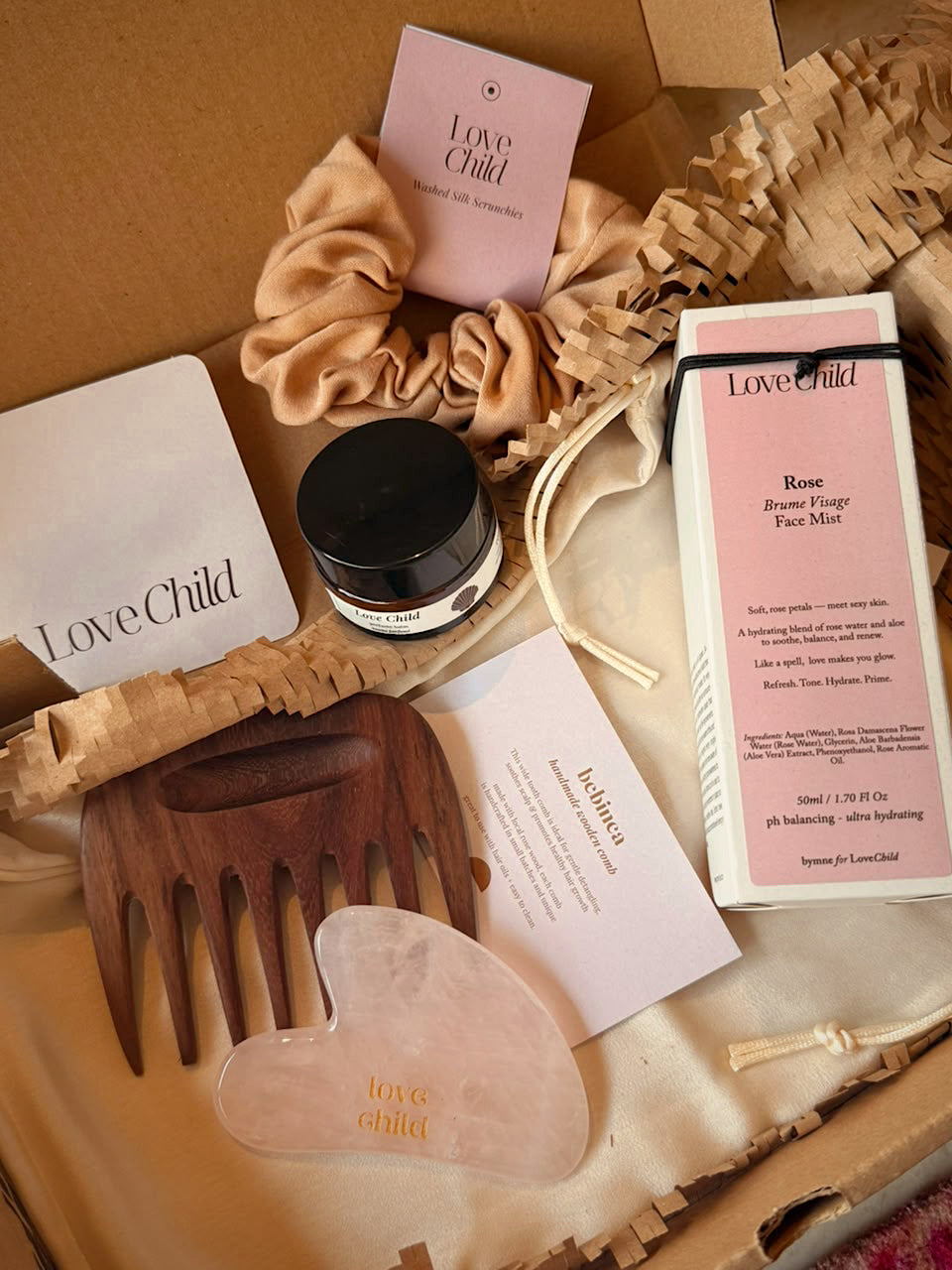 A curated beauty gift set from Love Child arranged in a kraft paper box. The set includes a rose facial mask box, a wooden hair extension comb, a translucent jade roller, a silk scrunchie, a small jar, and branded cards and packaging.