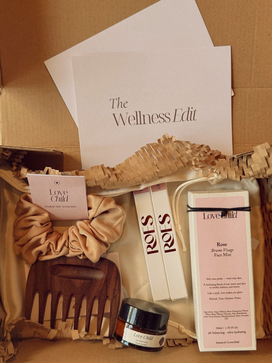 A curated wellness gift box from Love & Child, featuring a rose hair mask, a wooden comb, a satin scrunchie, and a small jar, arranged in a cardboard box and a card with details - 'The Wellness Edit'.