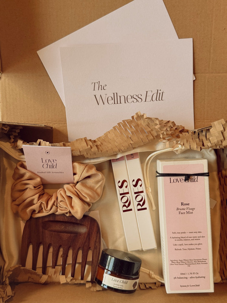 A curated wellness gift box from Love & Child, featuring a rose hair mask, a wooden comb, a satin scrunchie, and a small jar, arranged in a cardboard box and a card with details - 'The Wellness Edit'.