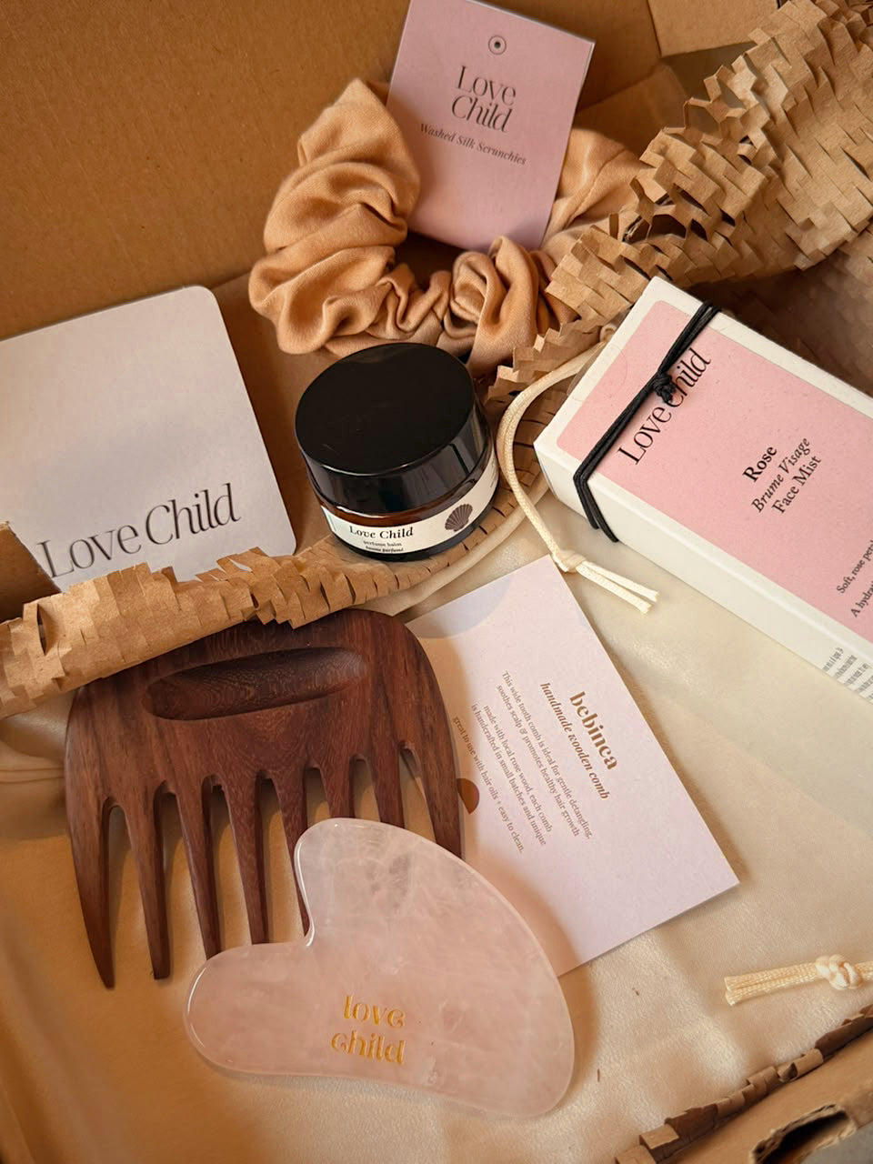 A curated beauty gift set from Love Child   featuring a handmade wooden hair gua sha comb, crystal face and body gua sha, washed silk scrunchie, face mist with rosewater, aloe and Vitamin E, perfume balm with coconut, rose and vanilla, and item details in a card.